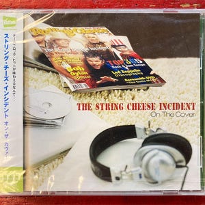 May include: A CD case featuring "The String Cheese Incident" album, with a magazine cover visible. The magazine has the text "TOP 365 Rock & Jam Songs" and "Bob Dylan Special". A pair of headphones is in the foreground.