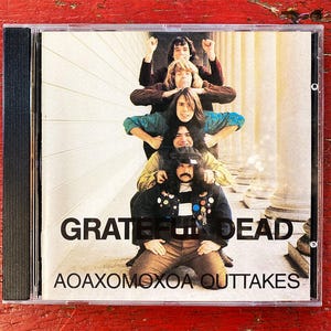 May include: A Grateful Dead CD case featuring a group of six men stacked on top of each other. The album title "AOAXOMOXOA OUTTAKES" is printed below the band's name. The background includes white columns.