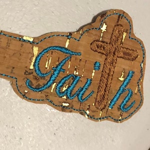 May include: A cork-backed key chain with a brown cross and the word "Faith" embroidered in turquoise blue.