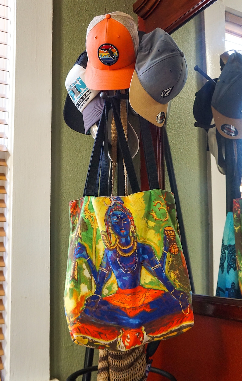 May include: A tote bag with a colorful image of a Hindu deity, Shiva, sitting in a meditative pose. The bag is hanging on a rack with several baseball caps.
