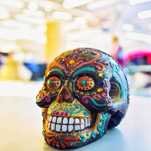 May include: A colorful, hand-painted sugar skull with intricate details and gold accents. The skull is black with bright colors like pink, blue, green, and yellow.