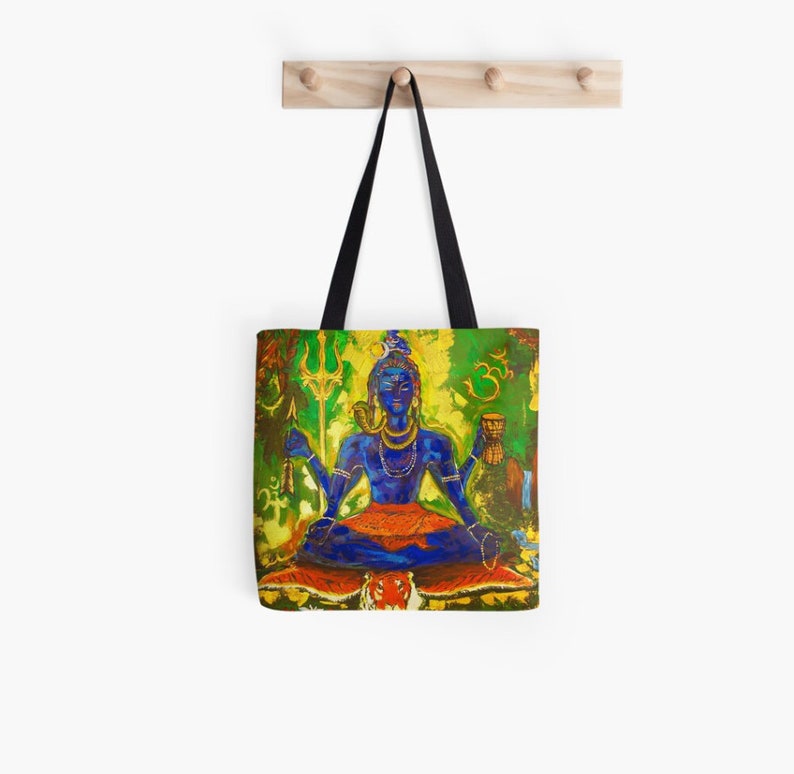 May include: A tote bag featuring a colorful painting of Shiva, a Hindu deity, sitting in a meditative pose on a tiger skin. He is holding a trident and a cup, and is surrounded by green and yellow foliage. The background includes the Om symbol and other Hindu symbols.