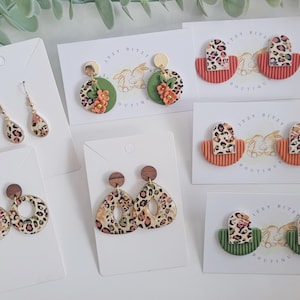 May include: A selection of handmade earrings in diverse designs. Styles include teardrop, round, and geometric shapes, all featuring a leopard print pattern. Some earrings have wooden accents, floral details, and gold-coloured posts. The earrings are displayed on white cards.
