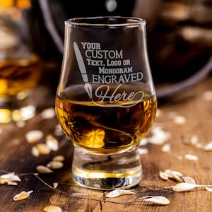 May include: A clear, tulip-shaped whiskey glass filled with amber liquid. The glass is engraved with the words "YOUR CUSTOM TEXT, LOGO OR MONOGRAM ENGRAVED Here". The glass sits on a wooden surface, with scattered grains and a blurred background.