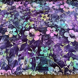 May include: A purple and blue quilted throw blanket with a floral pattern. The blanket has a frayed edge and is made of soft fabric.