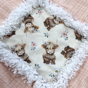 May include: A square, cream-colored baby blanket with a white, fluffy fringe. The center features a pattern of Highland cows with floral crowns and sprigs of flowers. The blanket is set on a pink, patterned surface.