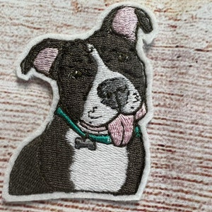 May include: Embroidered patch of a black and white dog with a pink nose and tongue sticking out. The dog is wearing a teal collar with a bone charm.