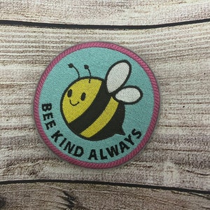 May include: A round, blue and pink embroidered patch with a cartoon bee and the text "Bee Kind Always".