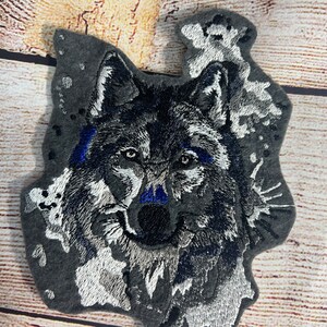May include: Embroidered gray wolf patch with blue accents. The wolf's face is detailed with white and black embroidery. The patch is surrounded by a white and gray watercolor-like design.