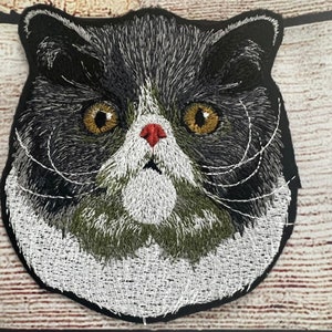 Könnte beinhalten: Gestickter Aufnäher eines schwarz-weißen Katers mit grünem und grauem Fell. Die Katze hat einen weißen Fleck auf der Nase und große, runde Augen.