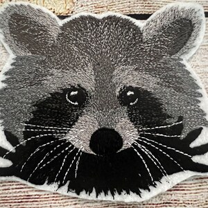 Könnte beinhalten: Gestickter Waschbär-Gesichts-Patch mit schwarzem und grauem Fell, schwarzen Augen und einer schwarzen Nase. Der Waschbär schaut direkt auf den Betrachter.