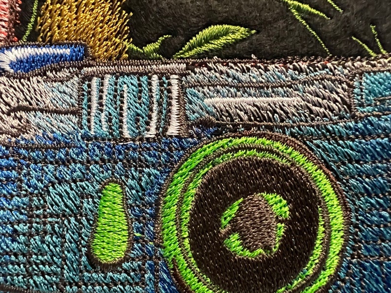 Peut inclure: Gros plan sur un appareil photo brod&eacute; bleu avec un objectif vert. L'appareil photo a un d&eacute;tail ray&eacute; noir et blanc sur le dessus.