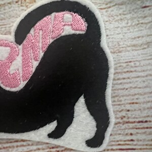 May include: A black cat-shaped patch with the word "DRAMA" embroidered in pink. The patch has a white border and is likely made of felt or a similar fabric. The cat's tail is raised, and the design is on a light-coloured wooden surface.