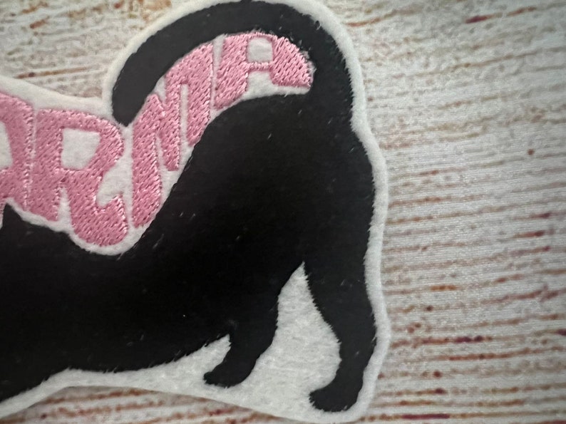 May include: A black cat-shaped patch with the word "DRAMA" embroidered in pink. The patch has a white border and is likely made of felt or a similar fabric. The cat's tail is raised, and the design is on a light-coloured wooden surface.