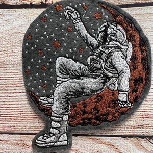 May include: Embroidered patch of an astronaut sitting on a crescent moon with stars in the background. The astronaut is wearing a white spacesuit and is reaching up to touch a star. The moon is brown and the background is gray.