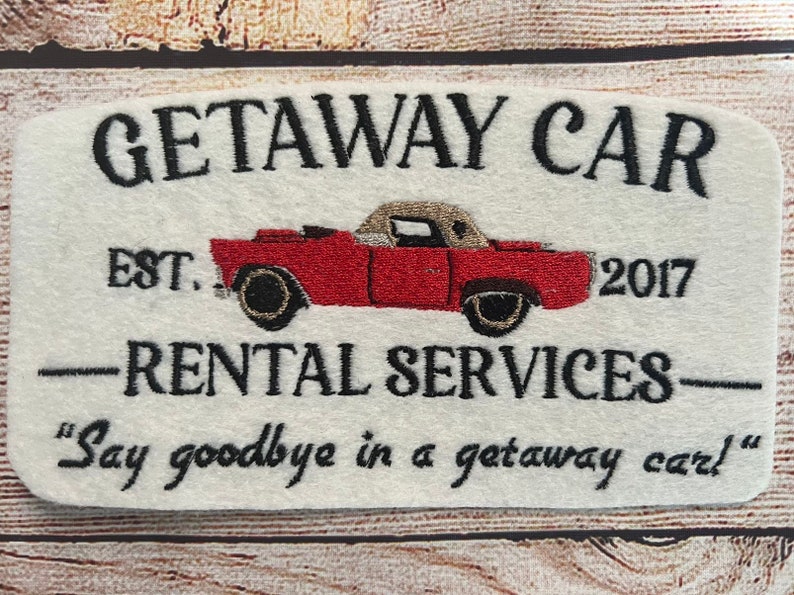 Puede incluir: Un cartel blanco con texto bordado en negro que dice "GETAWAY CAR EST. 2017 -RENTAL SERVICES- "Say goodbye in a getaway car!" Un coche rojo vintage con capo beige est&aacute; bordado en el centro del cartel.