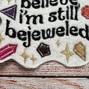 Puede incluir: Parche bordado con el texto "I believe I'm still bejeweled" en hilo negro. El parche es blanco con acentos de purpurina dorada y morada y presenta varias piedras preciosas de colores.