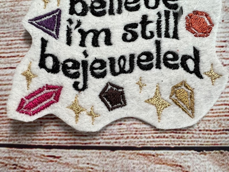 Puede incluir: Parche bordado con el texto "I believe I'm still bejeweled" en hilo negro. El parche es blanco con acentos de purpurina dorada y morada y presenta varias piedras preciosas de colores.