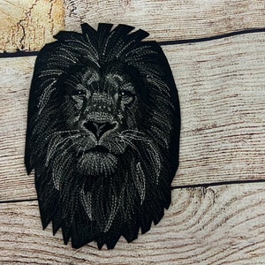 Puede incluir: Un parche de cabeza de león bordada en negro. El parche presenta costuras detalladas, mostrando la melena y los rasgos faciales del león. El parche está sobre un fondo claro de vetas de madera.