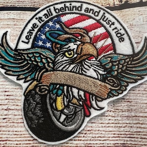 May include: Embroidered patch with an eagle holding a banner that reads "Leave it all behind and just ride". The eagle is in front of an American flag. The patch is white with gold, red, blue, and black embroidery.
