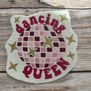 Pu&ograve; includere: Un cerotto in feltro bianco con ricami rosa e dorati. Il cerotto presenta una palla da discoteca, stelle e il testo "dancing queen".