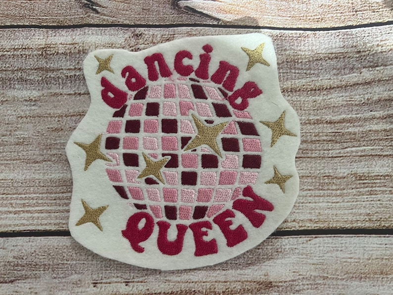Pu&ograve; includere: Un cerotto in feltro bianco con ricami rosa e dorati. Il cerotto presenta una palla da discoteca, stelle e il testo "dancing queen".