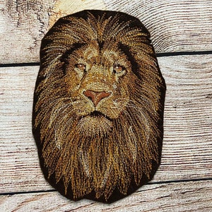 Puede incluir: Parche bordado de cabeza de león en tonos marrones y dorados. El león tiene una expresión seria y una melena llena.