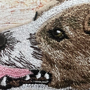 May include: Embroidered patch featuring a close-up of a dog's face. The dog has brown and white fur, a black nose, and a pink tongue. The patch is detailed with various thread colors, creating a textured appearance.