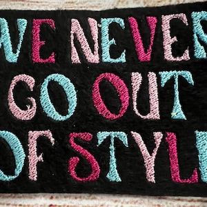 May include: Black embroidered patch with the text "WE NEVER GO OUT OF STYLE" in pink, blue, and white thread.