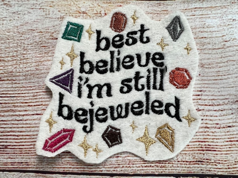 Puede incluir: Parche de fieltro blanco con el texto "best believe i'm still bejeweled" en bordado negro. El parche est&aacute; decorado con estrellas doradas brillantes y piedras preciosas bordadas de colores.