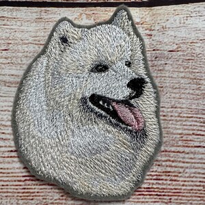 May include: Embroidered patch of a white Samoyed dog with a pink tongue sticking out. The dog is looking to the right and has a fluffy white coat.
