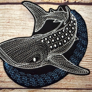 May include: Embroidered patch featuring a whale shark design. The whale shark is black with white stitching and white dot details. The patch is on a black background with blue stitching, set against a wood grain surface.