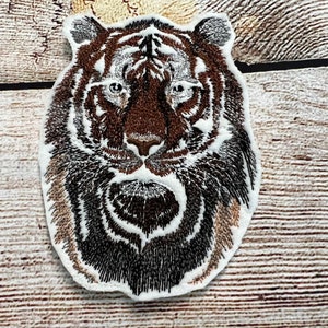 May include: An embroidered iron-on patch featuring a tiger's face. The tiger has black and brown stripes and a white background. The patch is ideal for adding a touch of wildness to your clothing or accessories.
