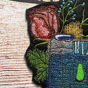 Peut inclure: Patch brod&eacute; repr&eacute;sentant un appareil photo vintage avec un objectif bleu et vert, entour&eacute; d'une rose rouge, de feuilles vertes et de fleurs bleues.