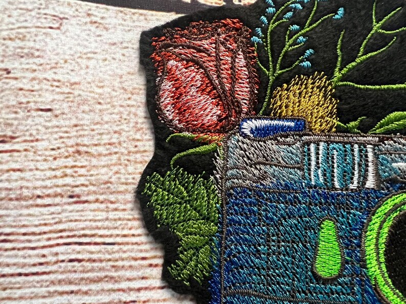 Peut inclure: Patch brod&eacute; repr&eacute;sentant un appareil photo vintage avec un objectif bleu et vert, entour&eacute; d'une rose rouge, de feuilles vertes et de fleurs bleues.