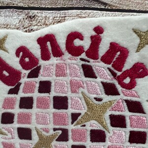 Pu&ograve; includere: Toppa ricamata con una palla da discoteca rosa e bordeaux e la parola "dancing" in lettere rosa e bordeaux. Ci sono cinque stelle dorate intorno alla palla da discoteca.