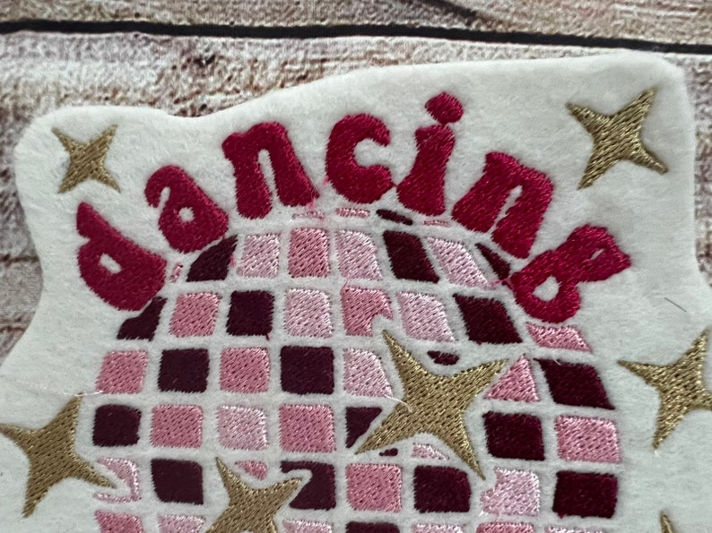 Pu&ograve; includere: Toppa ricamata con una palla da discoteca rosa e bordeaux e la parola "dancing" in lettere rosa e bordeaux. Ci sono cinque stelle dorate intorno alla palla da discoteca.