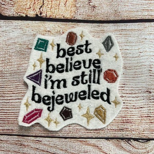 Puede incluir: Parche de fieltro blanco con texto bordado en negro que dice "best believe i'm still bejeweled". El parche est&aacute; decorado con piedras preciosas bordadas de colores y purpurina dorada.