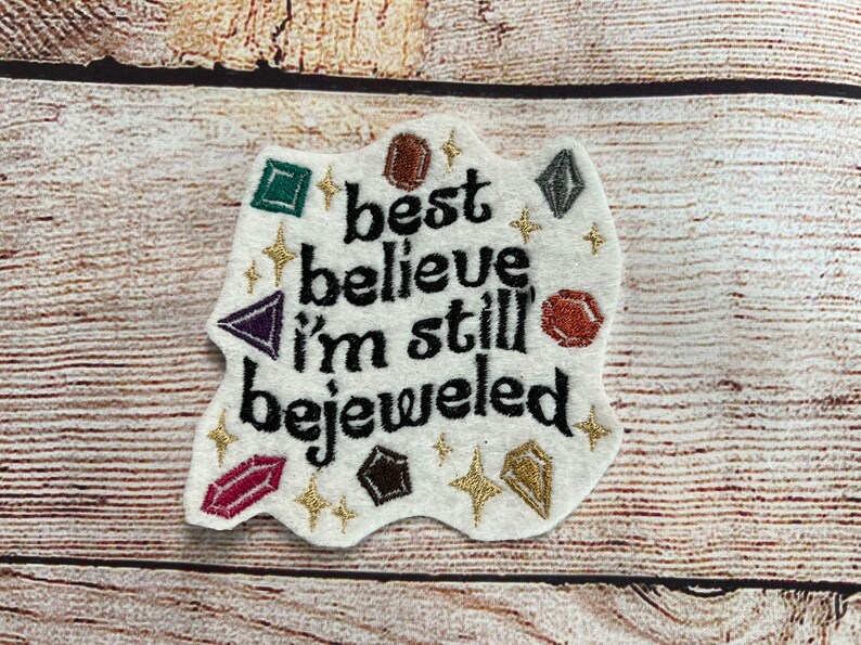 Puede incluir: Parche de fieltro blanco con texto bordado en negro que dice "best believe i'm still bejeweled". El parche est&aacute; decorado con piedras preciosas bordadas de colores y purpurina dorada.
