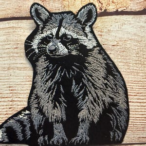 Puede incluir: Parche bordado con un diseño detallado de mapache. El parche es principalmente negro con acentos grises y blancos, mostrando el pelaje y los rasgos faciales del mapache. El mapache está representado en posición sentada.