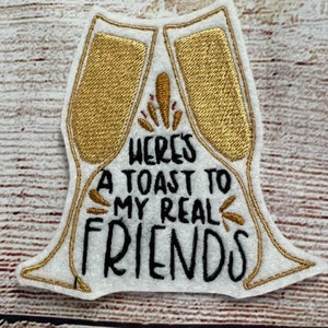 May include: Embroidered patch in the shape of two champagne flutes, with gold thread detailing. The patch reads "Here's a toast to my real friends" in black lettering. The patch is white with a gold border.