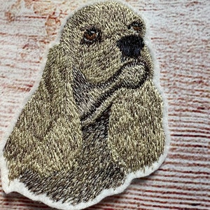 May include: Embroidered patch of a brown and black cocker spaniel dog with its head tilted to the side. The dog has a white patch on its chest and is looking to the right.