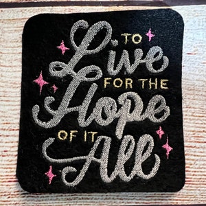 May include: Embroidered patch with a black background and silver lettering that reads "Live for the Hope of it All". The lettering is surrounded by pink stars.