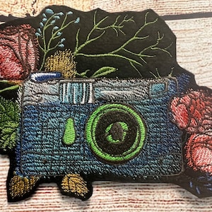 Peut inclure: Un patch brodé représentant un appareil photo bleu avec un objectif vert entouré de fleurs roses et vertes.