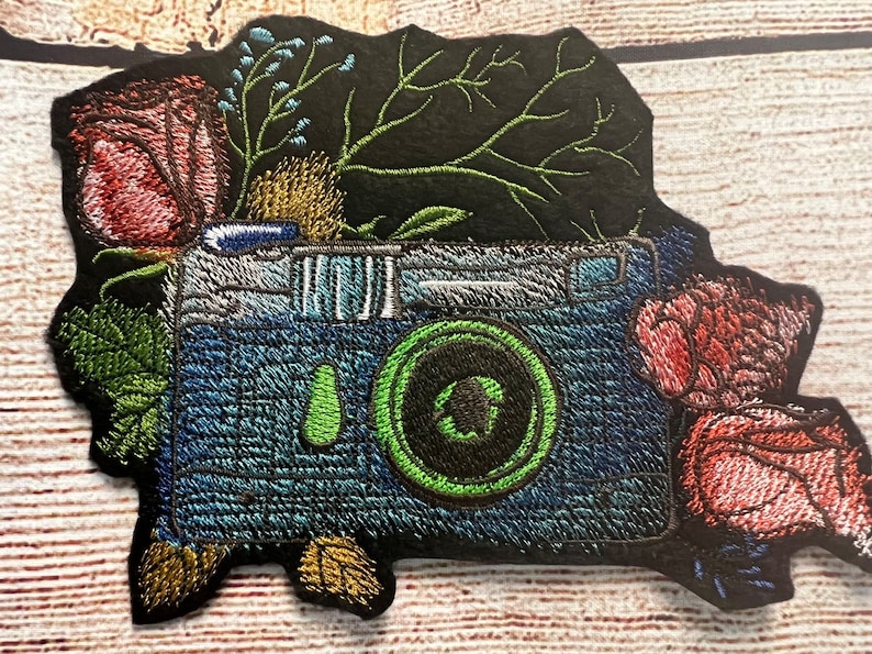 Peut inclure: Un patch brod&eacute; repr&eacute;sentant un appareil photo bleu avec un objectif vert entour&eacute; de fleurs roses et vertes.