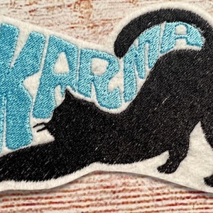 Op de afbeelding: Geborduurde patch met een zwarte kat die zich uitrekt, met het woord "KARMA" in lichtblauwe, gestileerde tekst. De patch heeft een witte rand, geschikt voor kleding of accessoires.
