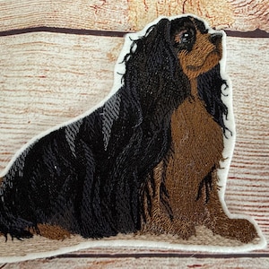 Op de afbeelding: Geborduurde patch met een Cavalier King Charles Spaniel hond. De patch toont de hond in een zittende houding, met zwarte en bruine vacht en een witte rand. De hond kijkt omhoog.