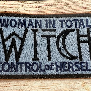May include: A blue embroidered patch with black stitching that reads "Woman in total control of herself" with the word "Witch" in a stylized font.