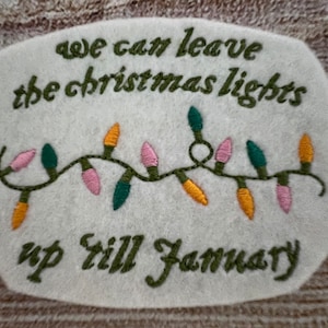 May include: White felt ornament with green embroidery that reads "we can leave the christmas lights up till january" and a string of colourful lights.