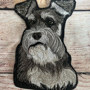 May include: Embroidered patch of a black and gray miniature schnauzer dog with a white beard. The dog is looking directly at the camera.